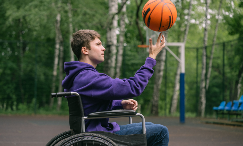 a young man in a manual wheelchair spins a basketball on his finger, outside at a court